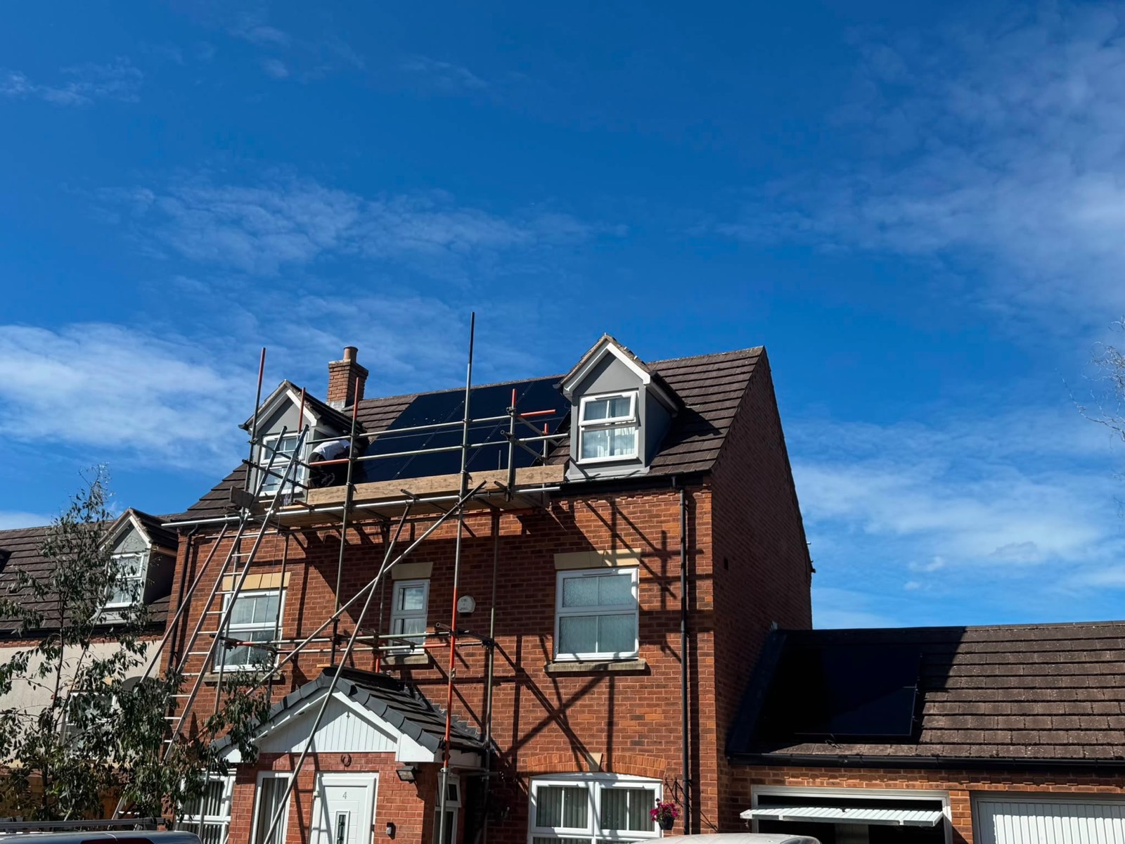 Solar panels being fitted to the roof of a three-storey brick house from scaffolding under a clear blue sky
